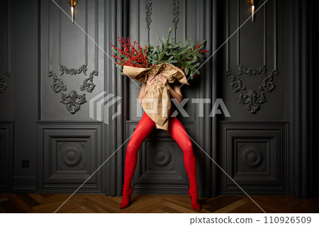 A long girl in red tights and high-heeled shoes holds a huge bouquet of fir branches and red berries wrapped in cardboard paper, she closes with a bouquet, photo studio with grey wall background 110926509