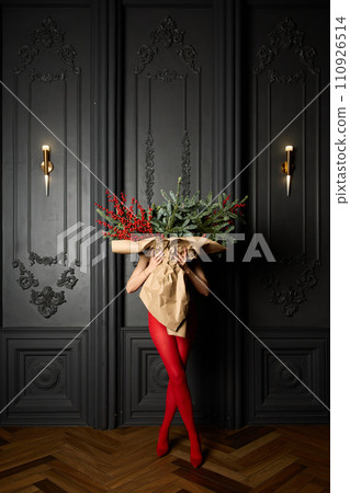 A long girl in red tights and high-heeled shoes holds a huge bouquet of fir branches and red berries wrapped in cardboard paper, she closes with a bouquet, photo studio with grey wall background 110926514