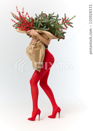 A long girl in red tights and high-heeled shoes holds a huge bouquet of fir branches and red berries wrapped in cardboard paper, she closes with a bouquet, photo studio with white background A long girl in red tights and high-heeled shoes holds a huge bouquet of fir branches and red berries wrapped in cardboard paper, she closes with a bouquet, photo studio with white background 110926531