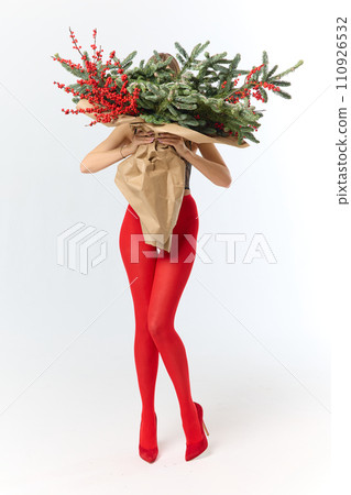 A long girl in red tights and high-heeled shoes holds a huge bouquet of fir branches and red berries wrapped in cardboard paper, she closes with a bouquet, photo studio with white background A long girl in red tights and high-heeled shoes holds a huge bouquet of fir branches and red berries wrapped in cardboard paper, she closes with a bouquet, photo studio with white background 110926532