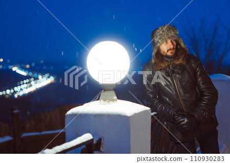 A man stands at a lantern at night in a hat with earflaps A man stands at a lantern at night in a hat with earflaps 110930283