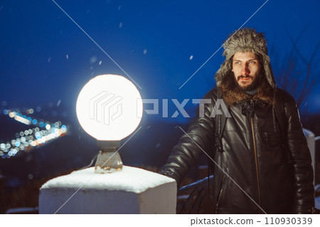 A man stands at a lantern at night in a hat with earflaps A man stands at a lantern at night in a hat with earflaps 110930339