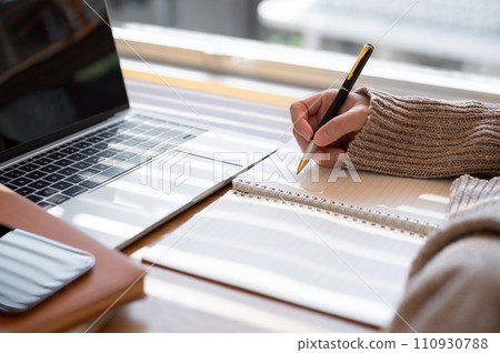 Close-up image of a woman is writing something in her notebook and working on her laptop indoors. Close-up image of a woman is writing something in her notebook and working on her laptop indoors. 110930788