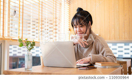 A positive, focused Asian woman is working on her assignment at a table indoors. A positive, focused Asian woman is working on her assignment at a table indoors. 110930964