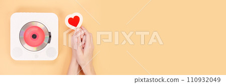 Banner of hands holding heart shape gift card next to white cd player with red disc on yellow background. Love music, Valentine day, Greeting, retro Love song, space for text. Banner of hands holding heart shape gift card next to white cd player with red disc on yellow background. Love music, Valentine day, Greeting, retro Love song, space for text. 110932049