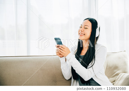 Attractive woman on sofa listens to music uses smartphone with headphones. Engaged in leisure relaxation and entertainment at home. Modern technology concept. 110932080