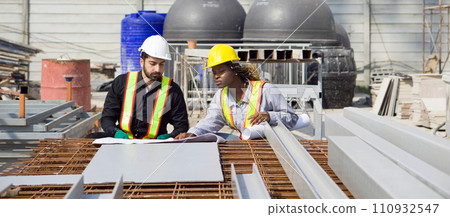 Two construction workers are working on construction tasks in front of a large building site with helmet and safety gear. 110932547