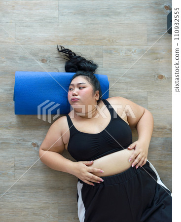 A plus-size woman lies down on a colorful yoga mat on the floor, relaxing after a soothing yoga session in a calm room. Top View 110932555