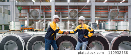 Two people shaking hands in front of large rolls of steel at an industrial facility. 110932588