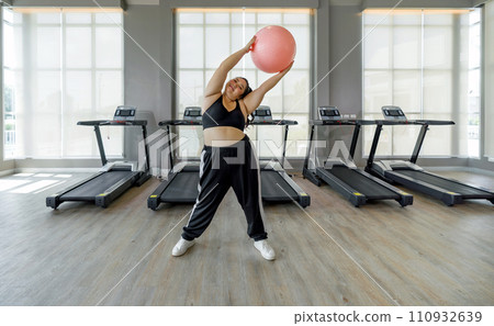 A woman is in a gym, gripping a bright pink ball with both hands, ready to exercise. A woman is in a gym, gripping a bright pink ball with both hands, ready to exercise. 110932639
