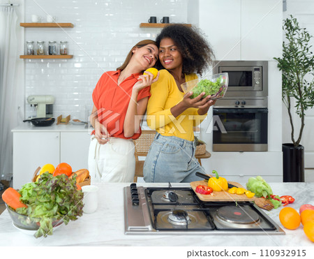 Two women stand in a kitchen holding various fruits and vegetables, smiling. 110932915