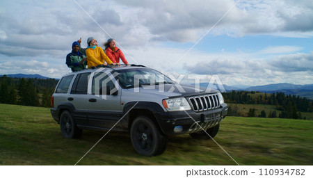 Hikers standing up through sunroof of car laugh and wave hands 110934782