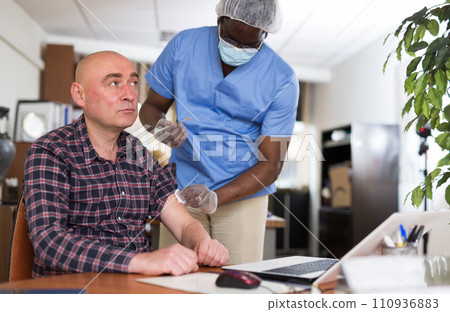 Qualified health worker in the protective mask vaccinates a male employee Qualified health worker in the protective mask vaccinates a male employee 110936883