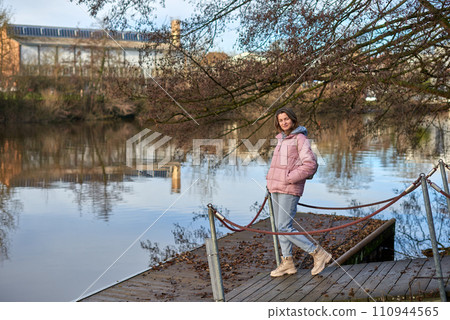 A young woman standing at the shore looking at the river in autumn sunny day. Street view, copy space for text, travel photo. Happy tourist woman on the bank of the river in autumn in warm clothes A young woman standing at the shore looking at the river in autumn sunny day. Street view, copy space for text, travel photo. Happy tourist woman on the bank of the river in autumn in warm clothes 110944565