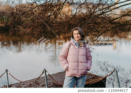A young woman standing at the shore looking at the river in autumn sunny day. Street view, copy space for text, travel photo. Happy tourist woman on the bank of the river in autumn in warm clothes A young woman standing at the shore looking at the river in autumn sunny day. Street view, copy space for text, travel photo. Happy tourist woman on the bank of the river in autumn in warm clothes 110944617