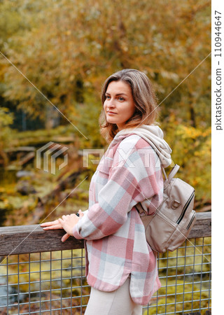 Portrait Of Cute Young Woman In Casual Wear In Autumn, Standing On Bridge Against Background Of An Autumn Park And River. Pretty Female Walking In Park In Golden Fall. Copy Space. Smiling Girl In The 110944647