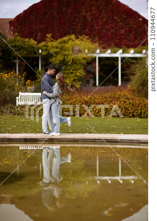 A couple in love hugs on the shore of a city pond in the European town. love story against the backdrop of autumn nature. Embraced by Love: Couple by the City Pond in a European Town. Autumn Affair 110944677