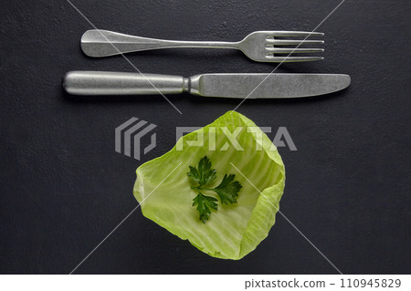 A leaf of white cabbage with a leaf of parsley and a fork with a knife on a black concrete background. Vegetarian nutrition concept for health and weight loss A leaf of white cabbage with a leaf of parsley and a fork with a knife on a black concrete background. Vegetarian nutrition concept for health and weight loss 110945829