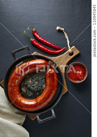 Ring of fried pork sausage on a black cast iron frying pan with hot pepper and ketchup on a black background. Top view with copy space Ring of fried pork sausage on a black cast iron frying pan with hot pepper and ketchup on a black background. Top view with copy space 110945845