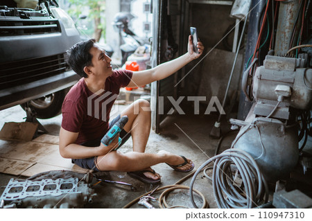 Young mechanic takes a selfie with cell phone while sitting with drill 110947310