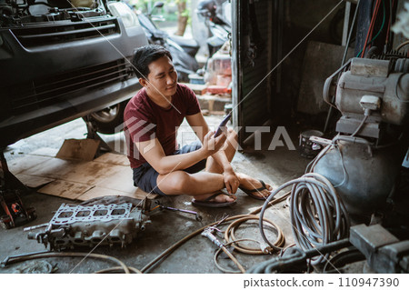 Young mechanic using a cell phone while sitting on the floor resting Young mechanic using a cell phone while sitting on the floor resting 110947390