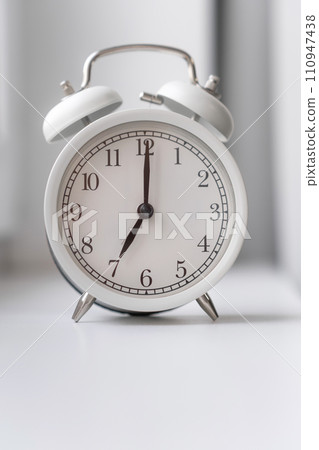 Close-up of a round white alarm clock on the windowsill in the bedroom. The hands on the clock show seven o'clock in the morning, time to get up. Retro alarm clock on the table. space for text Close-up of a round white alarm clock on the windowsill in the bedroom. The hands on the clock show seven o'clock in the morning, time to get up. Retro alarm clock on the table. space for text 110947438