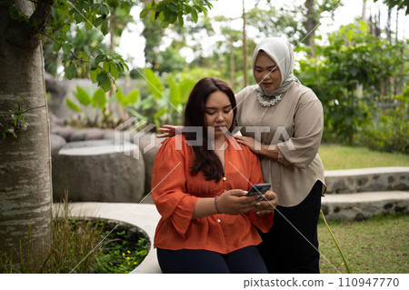 businesswoman holding phone sitting in park with hijab coworker comforting her 110947770