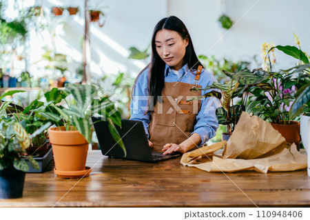 Successful Asian female florist wearing apron working laptop in own floral studio, check customers order in cozy modern interior full green plants. Plants business, start up. Small business, home Successful Asian female florist wearing apron working laptop in own floral studio, check customers order in cozy modern interior full green plants. Plants business, start up. Small business, home 110948406
