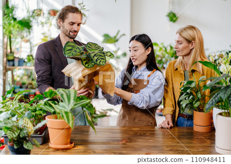Adult Asian woman worker gardener wearing apron packing in paper and selling house plant pots for happy European couple in flower shop. Plants business, start up. Florist handing over plants to male Adult Asian woman worker gardener wearing apron packing in paper and selling house plant pots for happy European couple in flower shop. Plants business, start up. Florist handing over plants to male 110948411