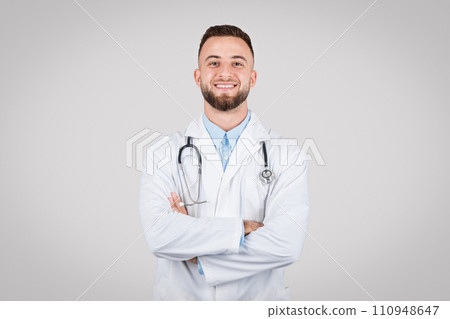 Confident young doctor posing with arms crossed and stethoscope around his neck against grey background 110948647