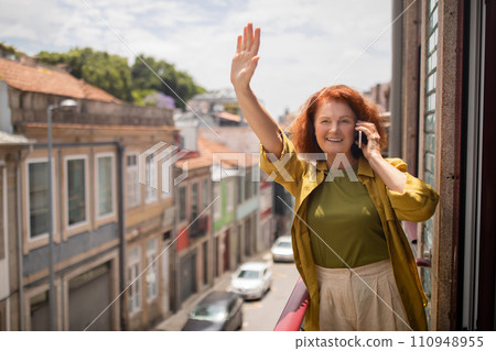 Cheerful senior woman standing on balcony, waving hand and talking on cellphone 110948955