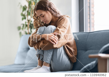 Distressed teenage girl sits on couch at home hugging knees Distressed teenage girl sits on couch at home hugging knees 110948962
