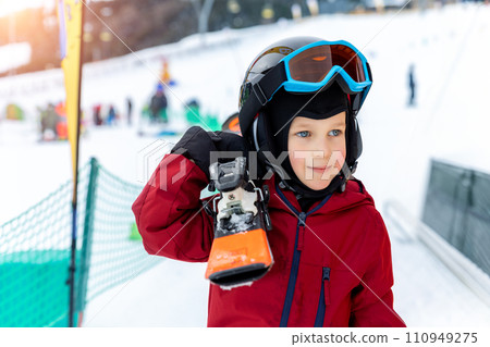 Cute adorable little kid boy enjoy having fun walking with ski on shoulder at alpine mountain skiing resort on bright winter day. Toddler beginner skier rest of training in ski school 110949275