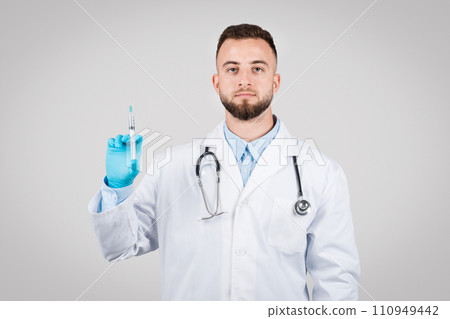 Serious young male doctor with syringe, white coat, gloves, grey background 110949442