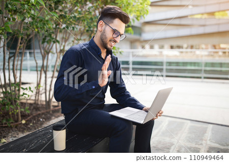 Cheerful arab businessman looking at laptop webcam waving hand outside Cheerful arab businessman looking at laptop webcam waving hand outside 110949464