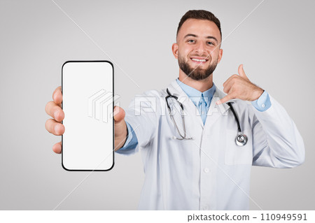 Young cheerful doctor showing phone with blank screen and gesturing call me Young cheerful doctor showing phone with blank screen and gesturing call me 110949591