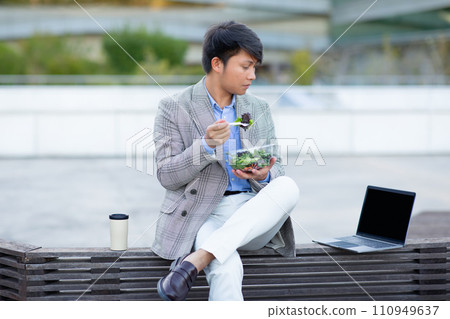 Asian businessman eating salad looking at laptop in urban area 110949637