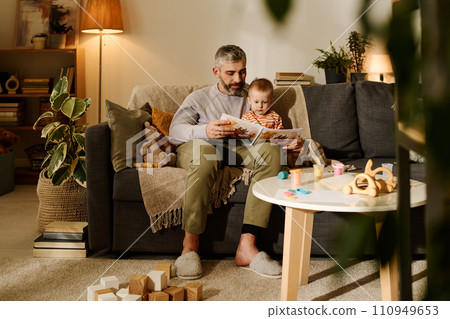 Reading book to baby son in living room 110949653