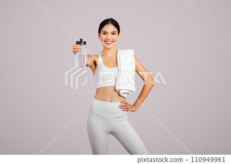 Smiling slim caucasian lady in sportswear with towel showing bottle of water, isolated on grey studio background Smiling slim caucasian lady in sportswear with towel showing bottle of water, isolated on grey studio background 110949961