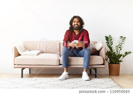 Positive indian man sitting on couch and using cell phone 110950083