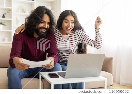 Emotional indian spouses sitting on couch, looking at laptop screen 110950092
