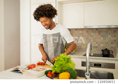 black guy peeking at recipes book chopping vegetables at kitchen black guy peeking at recipes book chopping vegetables at kitchen 110950651