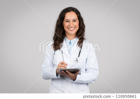 Positive woman doctor practitioner in white coat using digital tablet and smiling, grey background, studio shot Positive woman doctor practitioner in white coat using digital tablet and smiling, grey background, studio shot 110950661