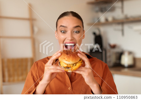 Joyful woman holding a juicy burger in kitchen Joyful woman holding a juicy burger in kitchen 110950863
