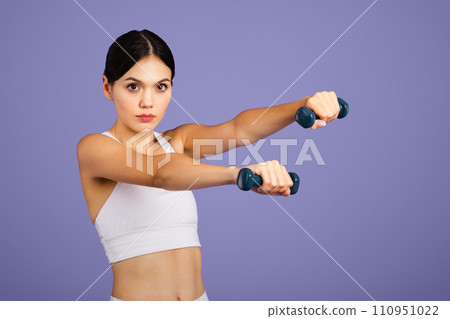 Serious caucasian slim woman in sportswear doing exercises with hand dumbbells, looking at camera, purple background Serious caucasian slim woman in sportswear doing exercises with hand dumbbells, looking at camera, purple background 110951022