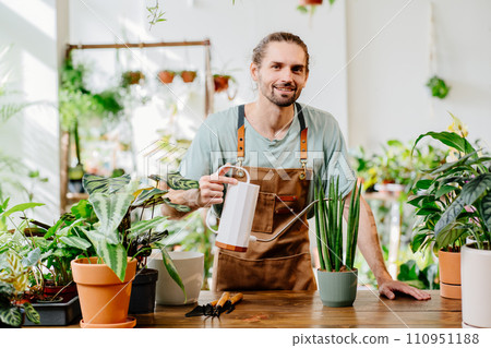 Young man wearing apron carefully watering potted houseplants at home indoors. Concept of plants care and home garden. 110951188