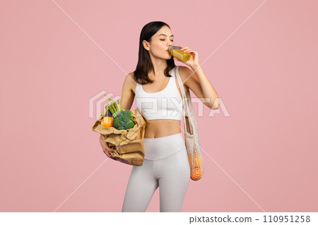 Sporty caucasian woman in sportswear sipping nutritious smoothie and holding bag of fresh vegetables, pink background 110951258
