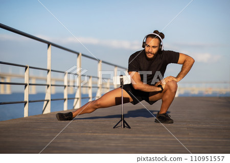 fitness guy in deep lunge, streaming exercise via smartphone outdoors 110951557