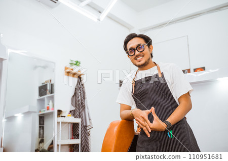 asian male hairstylist leaning on barber chair with smiling face 110951681