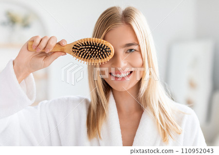Blonde woman in bathrobe playfully covers eye with hairbrush, indoor 110952043